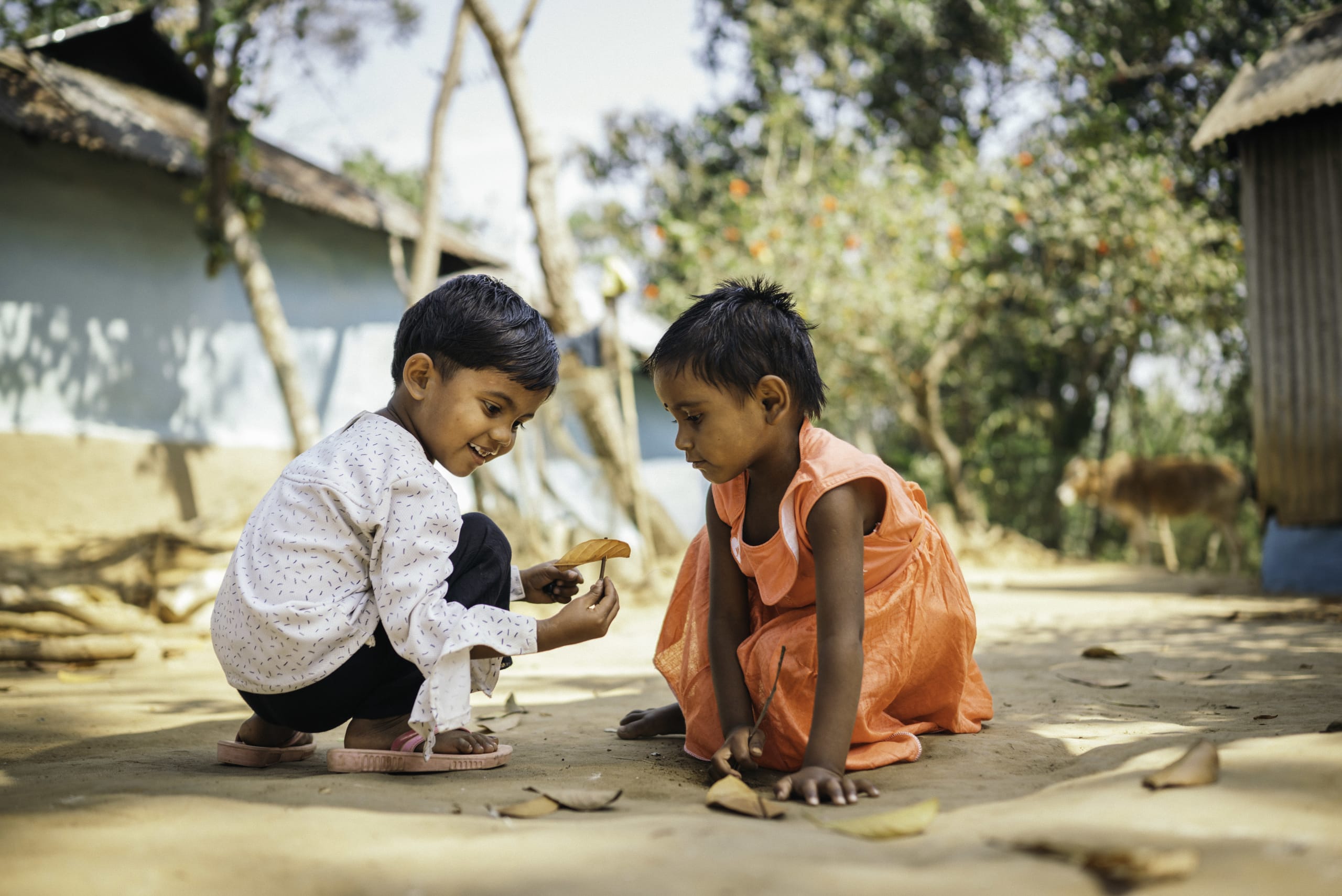 Two young children play with some fallen leaves on the ground outside.