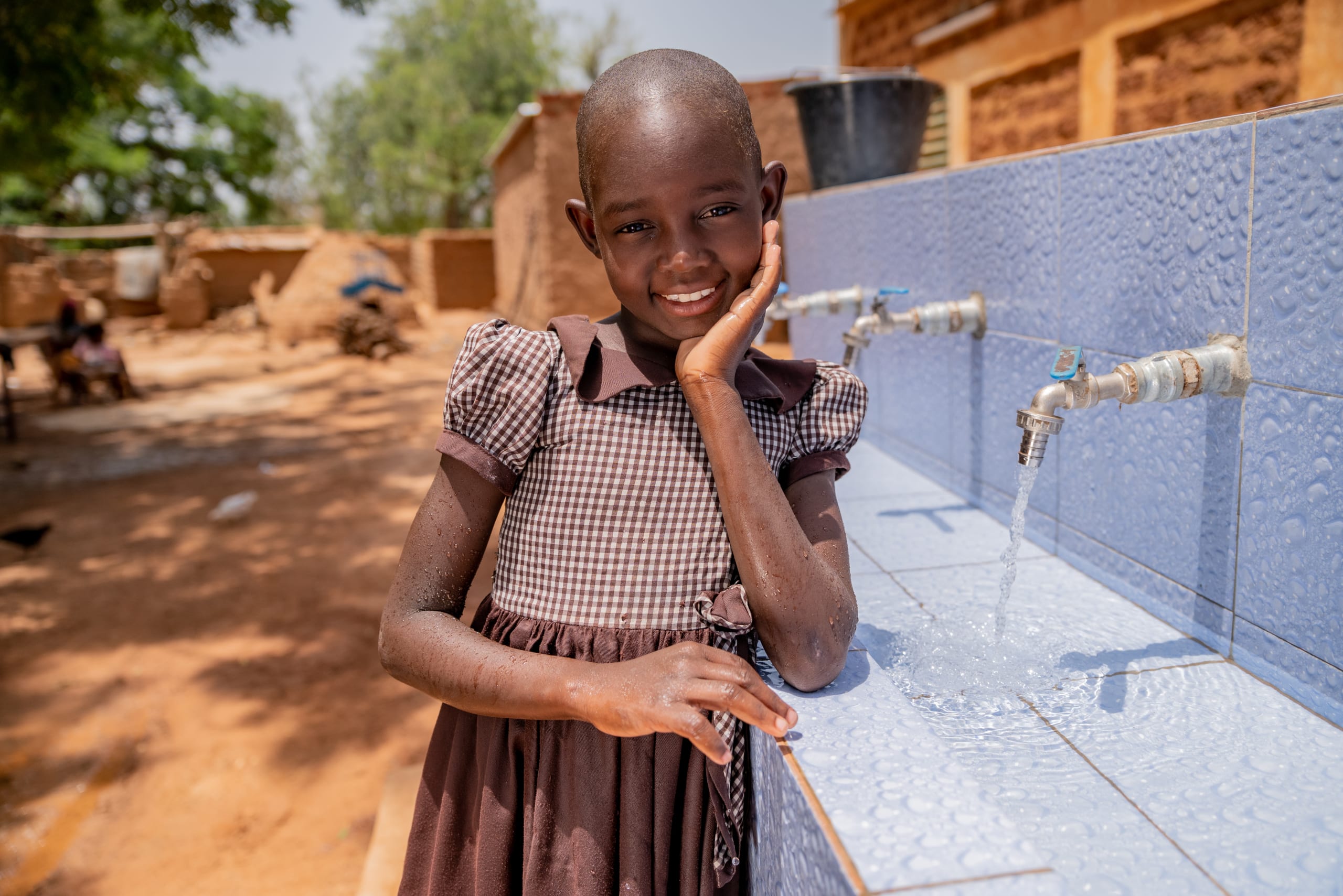 A young girl in a brown dress leans against a blue block of sinks with running water in the faucets