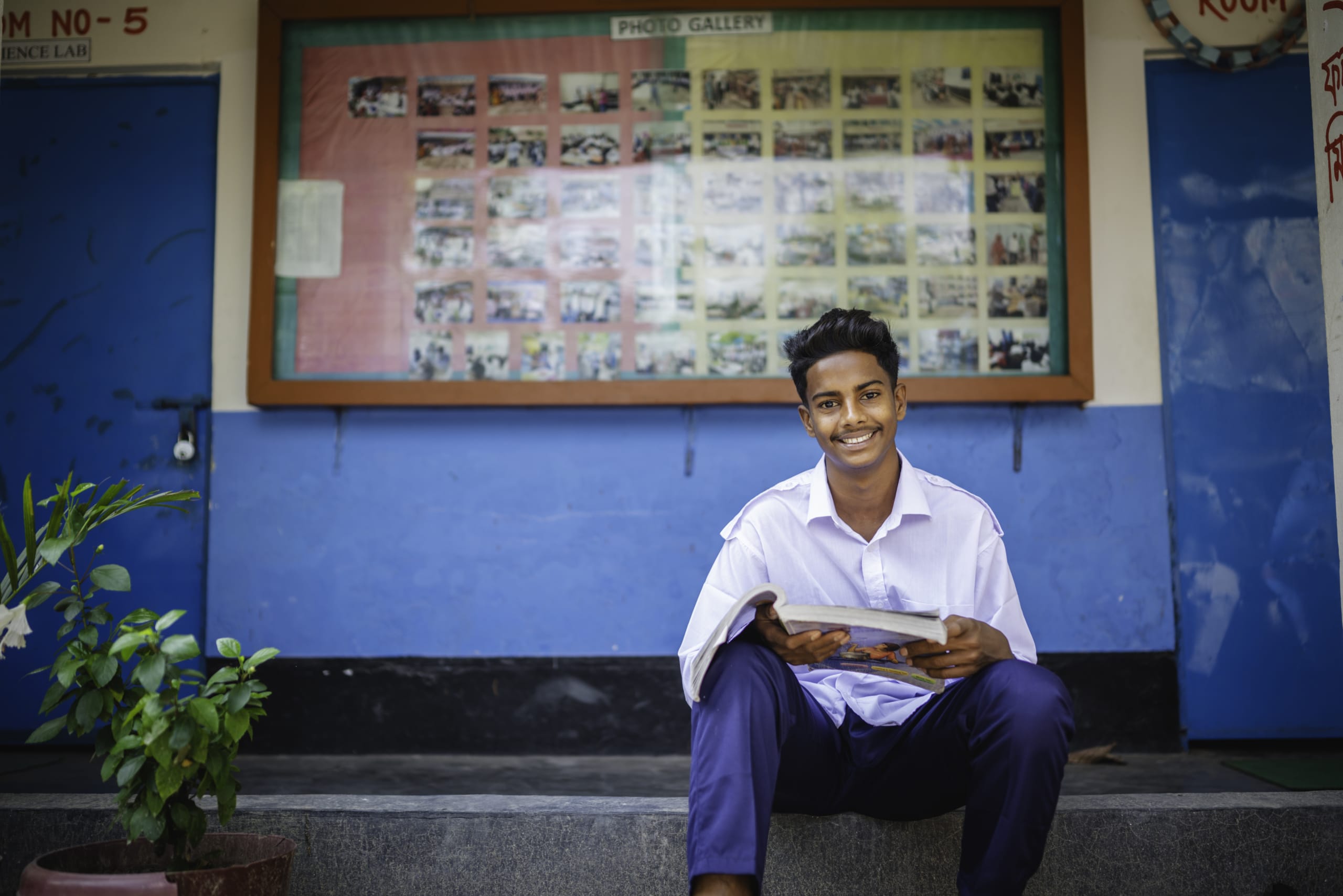 Shanto sits on a step in a white shirt and jeans. He is holding a textbook and smiling.