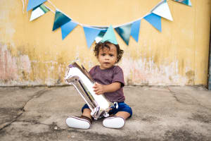 A little boy sits in front of a yellow wall with a "one" balloon. The wall has blue bunting on it.