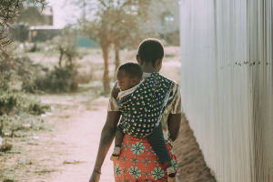 Woman carries baby on her back in a polka dot carrier. They are walking away from the camera.