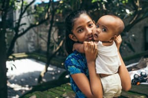 Girl in a blue dress is holding a baby and smiling. The baby is wearing a white shirt and they are in the trees.