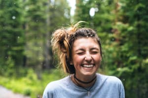 A girl in laughing with her eyes closed with her hair up in a bun. She has a blue t-shirt and 2 necklaces, and is surrounded by the forest. It is snowing