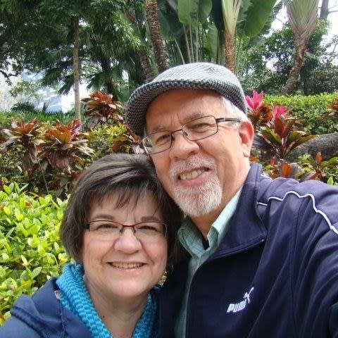 A woman with short brown hair stands beside a man with glasses and a hat. They both smile.