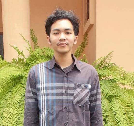 A man in a grey plaid shirt stands looking at the camera