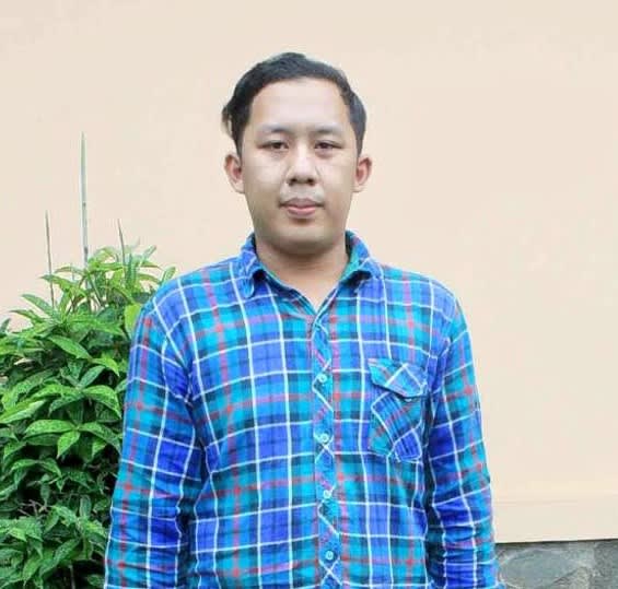 A man in a blue shirt stands looking at the camera