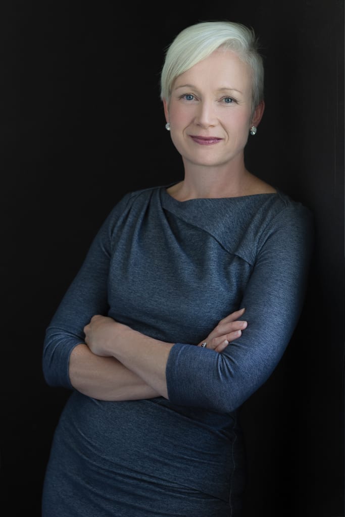Elegant woman stands in a casual but confident pose. She has short light hair and a blue dress. Kate Rock-Thompson from Rock Thompson Financial provides advice about year-end giving