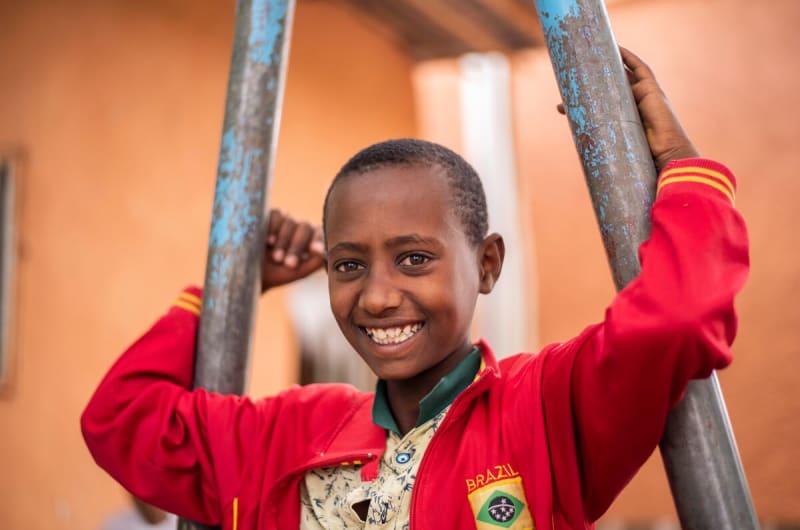 Boy in a red jacket smiles at the camera while holding onto a swing set.