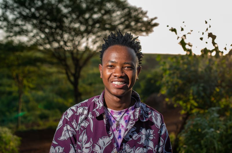 Arnold stands outside in front of a tree with a purple patterned shirt on.
