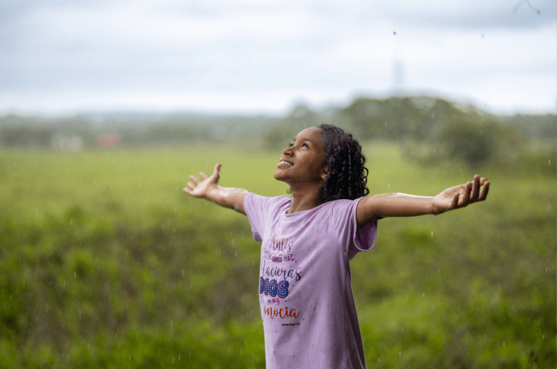 A little girl in a purple t-shirt stands in a field with her hands outstretched and looks to the sky.