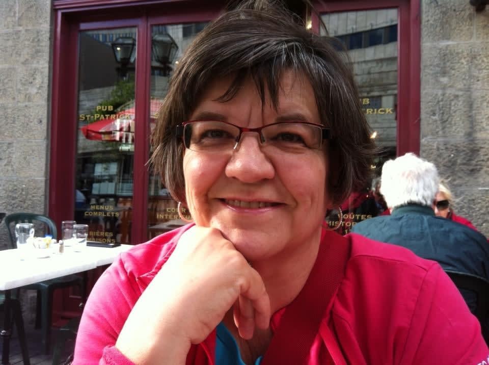 A woman with short brown hair wears a pink sweater and smiles at the camera with her chin resting on her hand.
