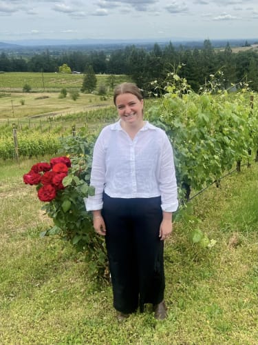 Tasting Room & Social Media Associate Helen Plotkin at Compris Vineyard with the beautiful valley view and town of Newberg in the background.