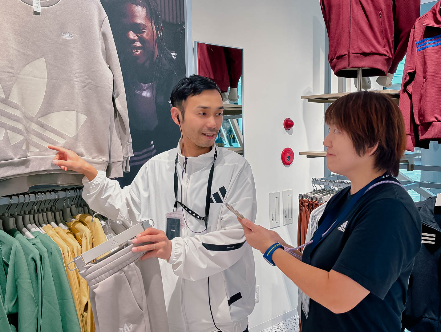 A man and a woman motion towards an adidas sweatshirt hanging on a rack and discuss it.