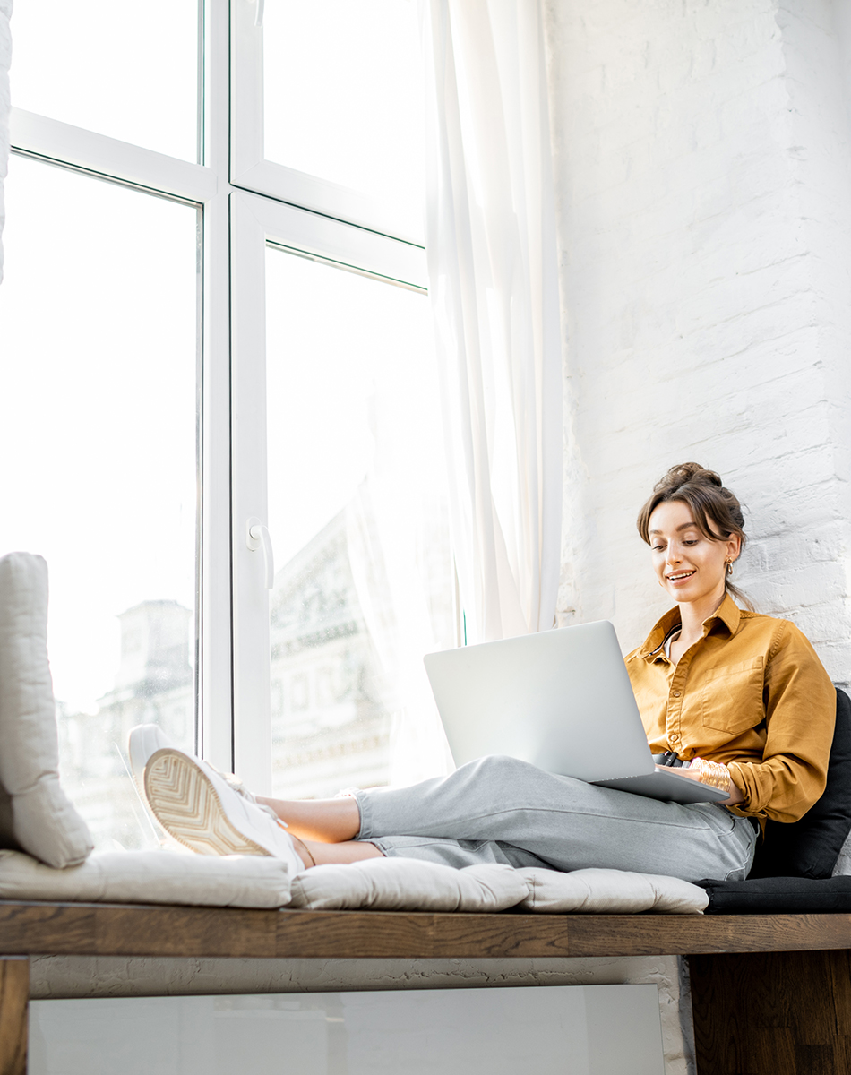 Woman sitting next to windowsill working on laptop