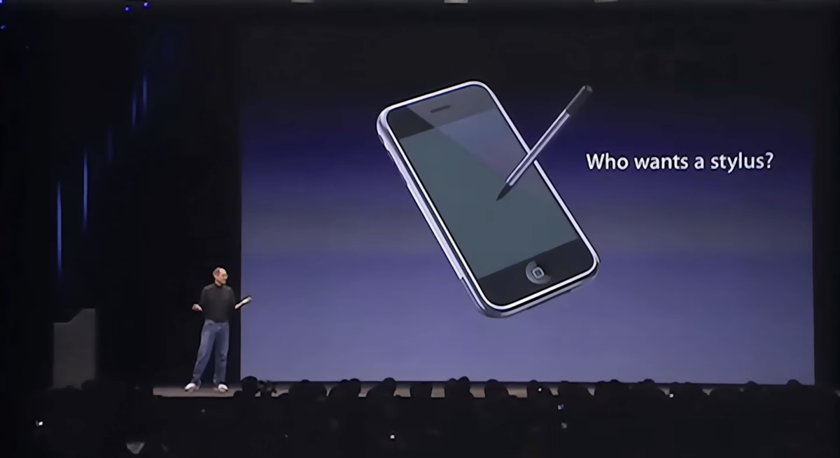 Steve Jobs on stage beside a slide of an iPhone with a stylus poised over it, captioned ’Who wants a stylus?’.