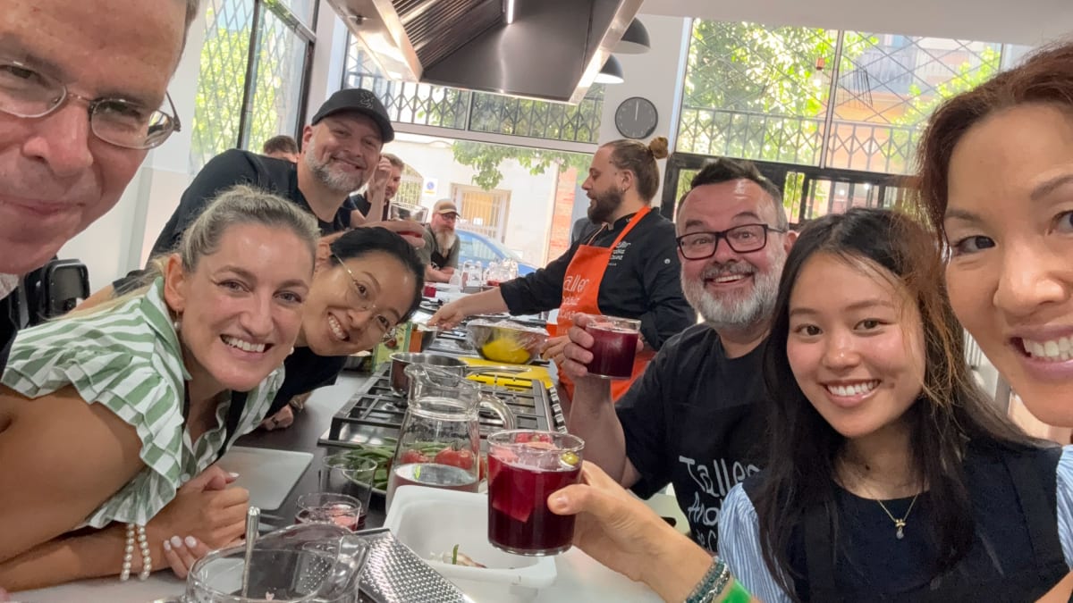 A group of smiling cooking class participants raising glasses of sangria at a communal table in a bright kitchen.