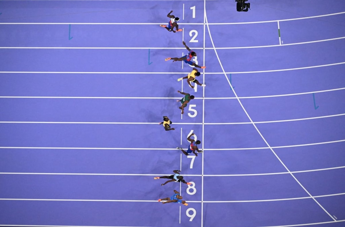 Overhead view of sprinters crossing the finish line in a 100-meter race on a blue track, with the athlete in lane 7 narrowly leading by a small margin.