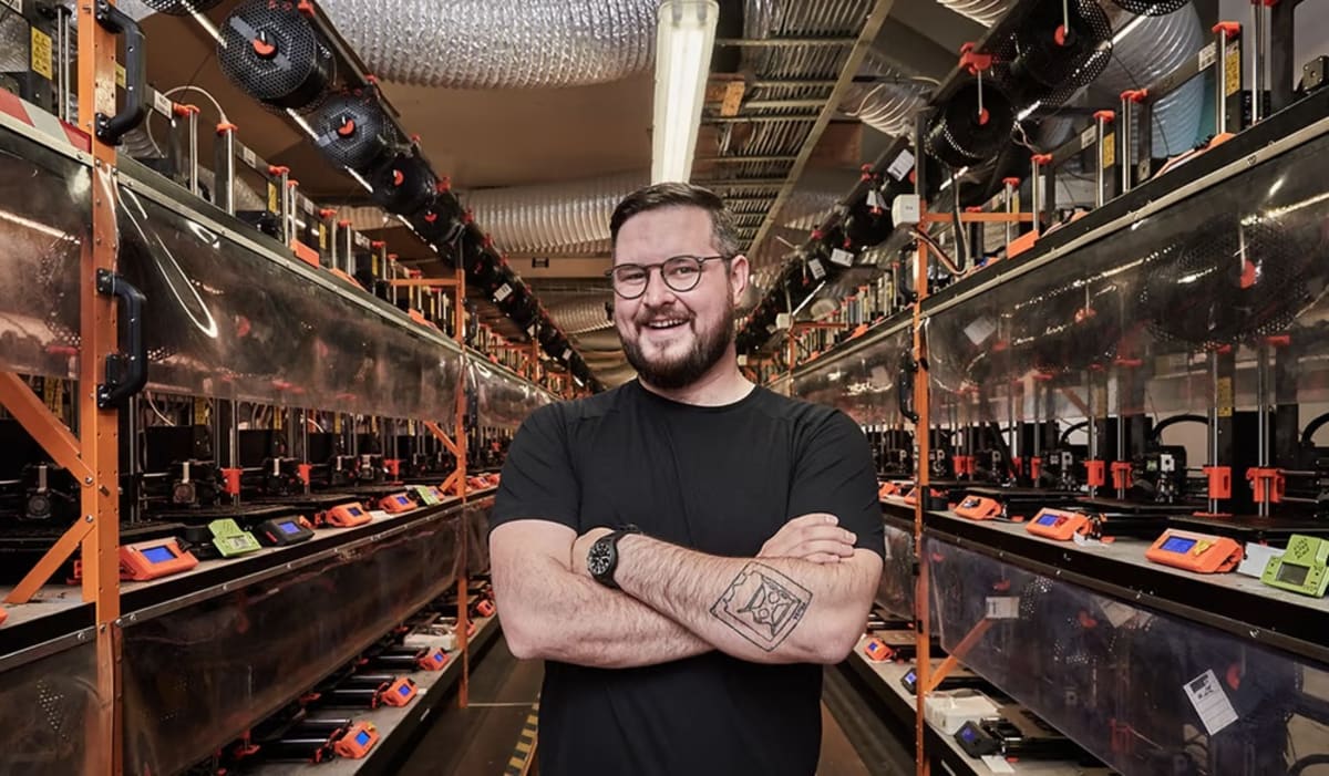Prusa&rsquo;s owner Josef Prusa smiling and standing in front of an inventory of 3D printers