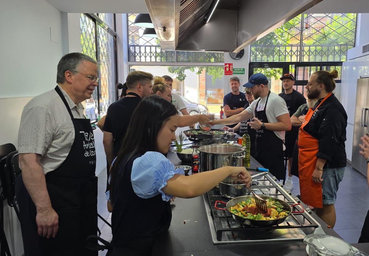 A cooking class in Seville with participants wearing black aprons gathered around a kitchen counter with knives, cutting boards, and glass pitchers.