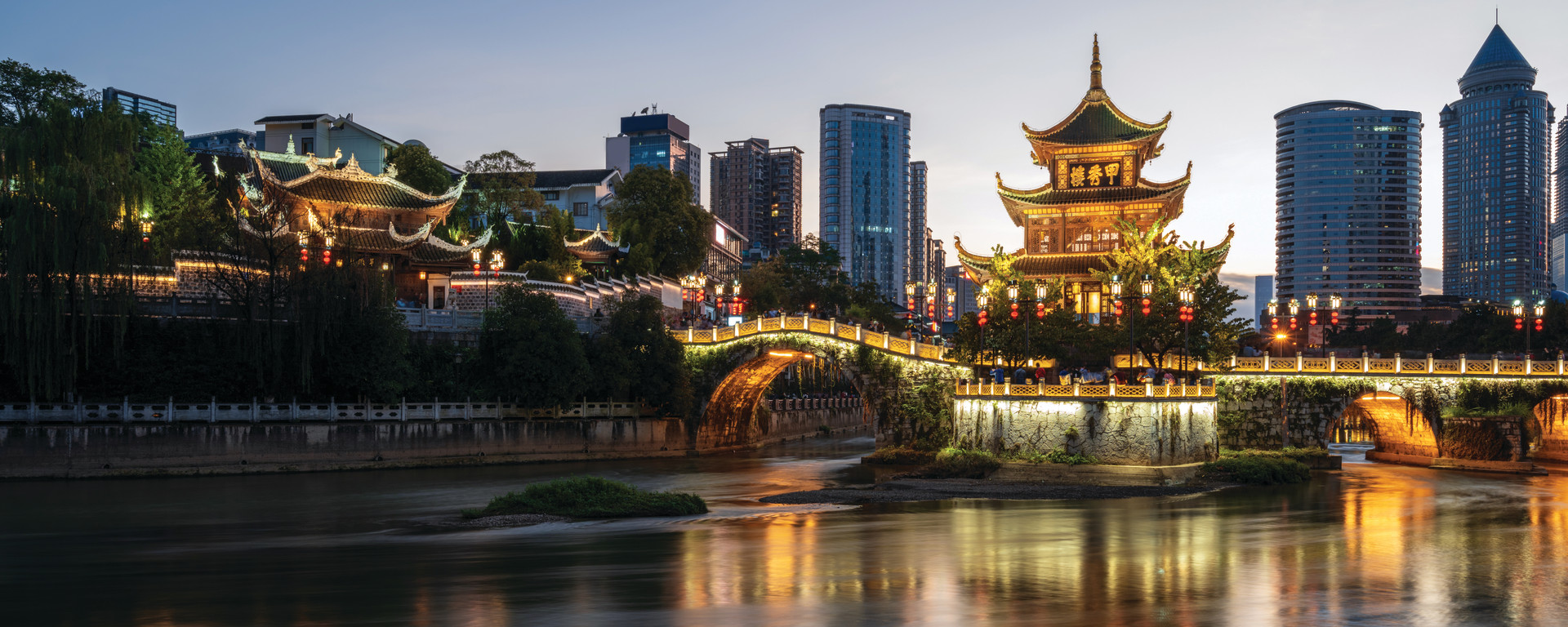 A vibrant Asian cityscape featuring a traditional temple perched above a river, overlooking the modern skyline. The scene blends ancient architecture with contemporary high-rise buildings, with the temple standing prominently against the backdrop of the city, creating a striking contrast between heritage and urban life.