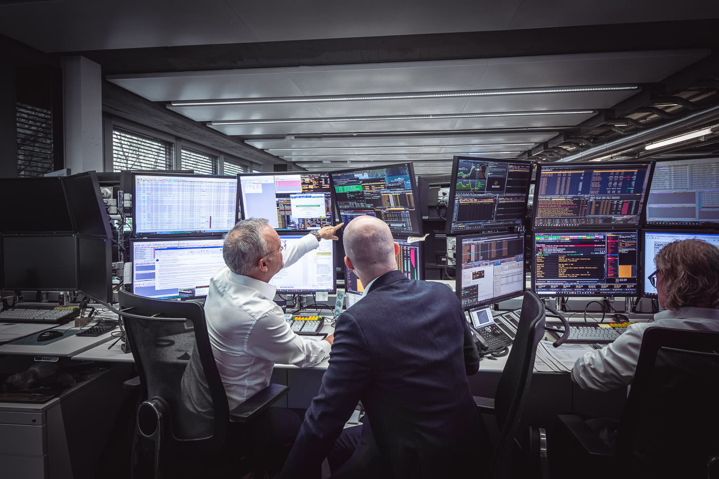 Drei Männer sitzen an Büroschreibtischen mit mehreren Computerbildschirmen für Finanzanalysen. Einer der Männer zeigt auf einen Bildschirm, um dem anderen Mann etwas zu erklären.