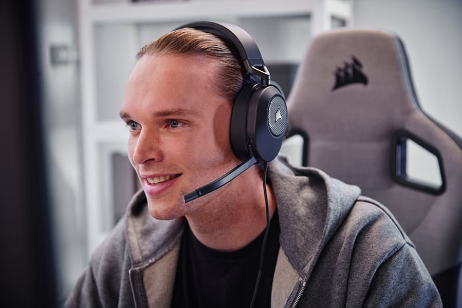 A smiling, man looking at a monitor while wearing a pair of CORSAIR HS65 SURROUND gaming headset in black sitting in a gaming chair.