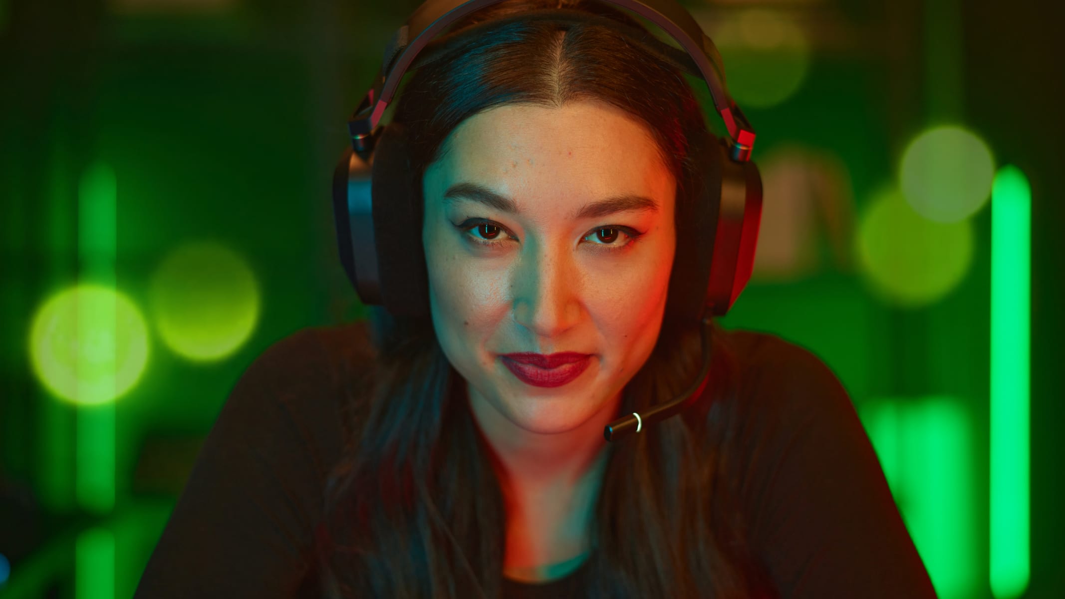 A smirking brunette female is wearing a black CORSAIR HS80 RGB WIRELESS gaming headset.