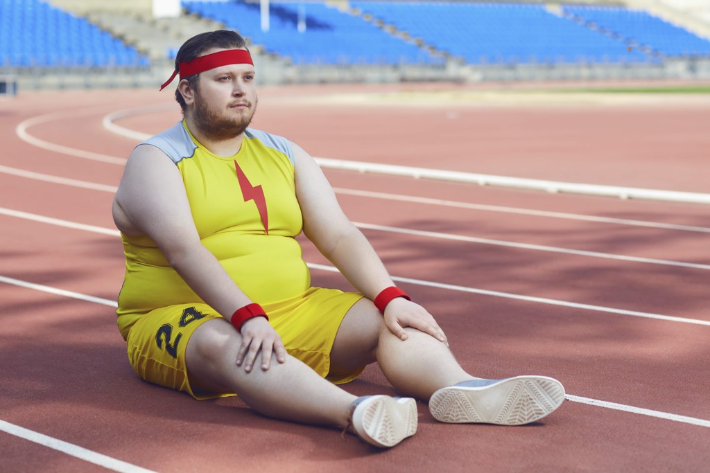 Ein fülliger Läufer in Supermankluft sitzt auf dem Tartanboden.