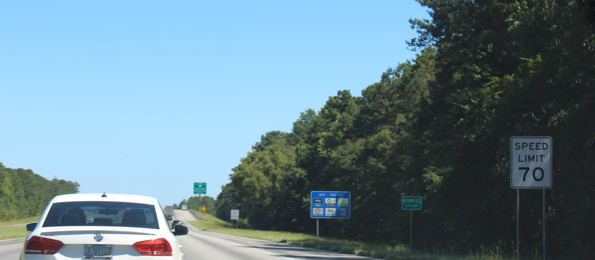 Autobahn mit einem weißen Auto und Straßenschildern am Waldrand.