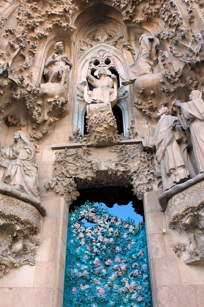 Outside the Sagrada Familia. The image was taken of the Nativity Facade on the east side of the basilica. The statues, including Joseph and the Virgin Mary are facing towards Jesus in the middle. At the bottom of the image is a panel with colourful green leaves, white and pink flowers, birds and nests that climb up the glass.