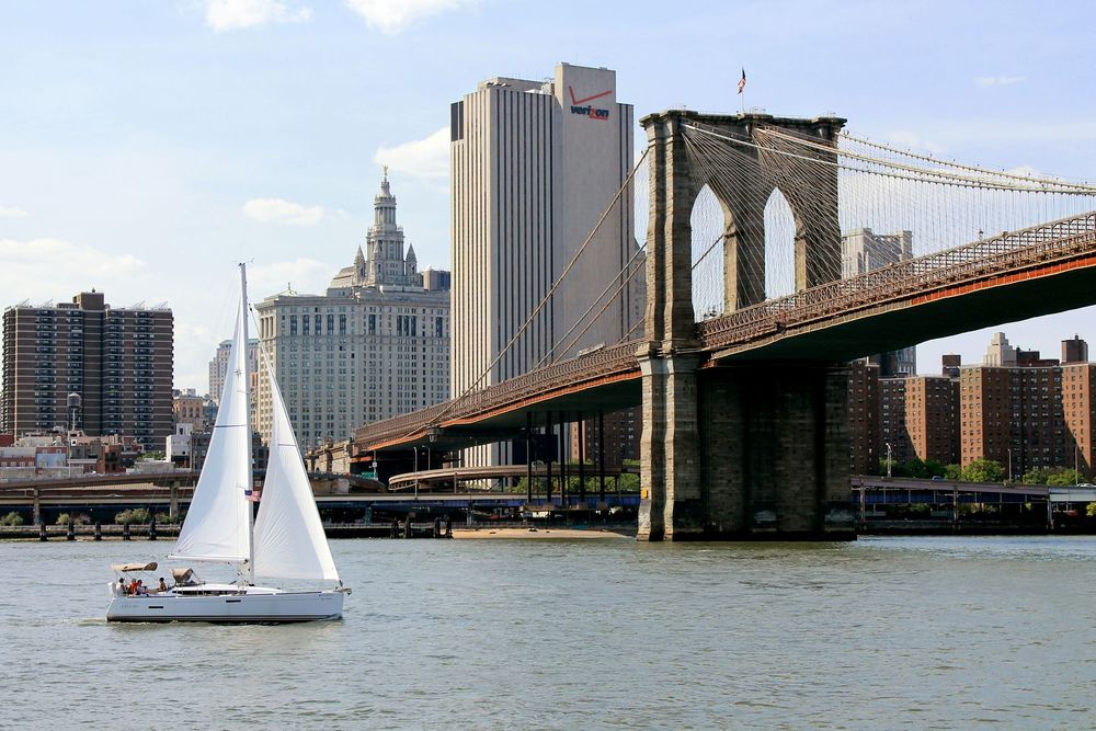The Brooklyn bridge as visible from the Brooklyn side of the river. A sailboat is passing through the image from the left side. The Verizon building is visible in the background.