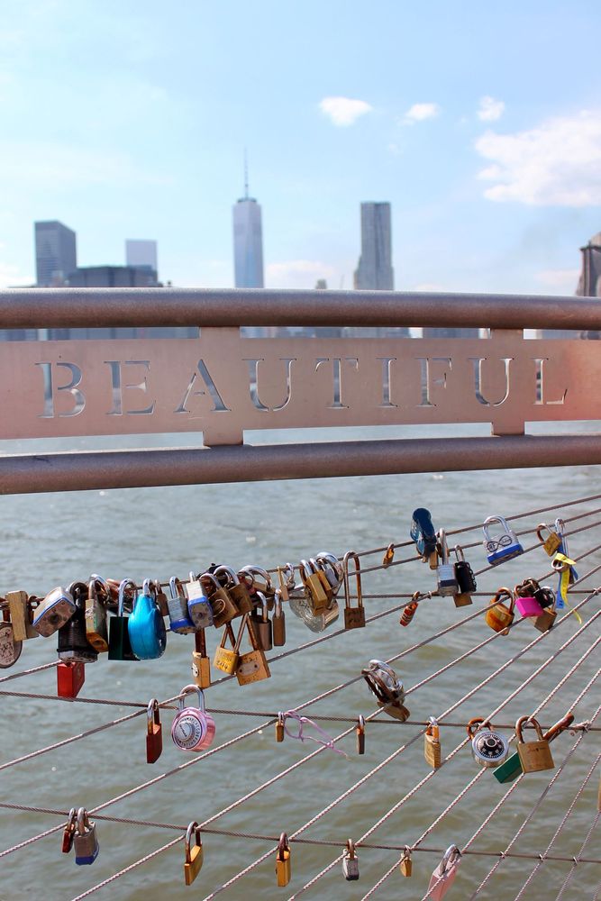 At Pier 1 in Brooklyn, New York. The New York City skyline is visible in the background. In the foreground is a closeup of a metal fence along the pier where people have attached locks. The fence reads Beautiful and the Hudson River is visible through the gaps.