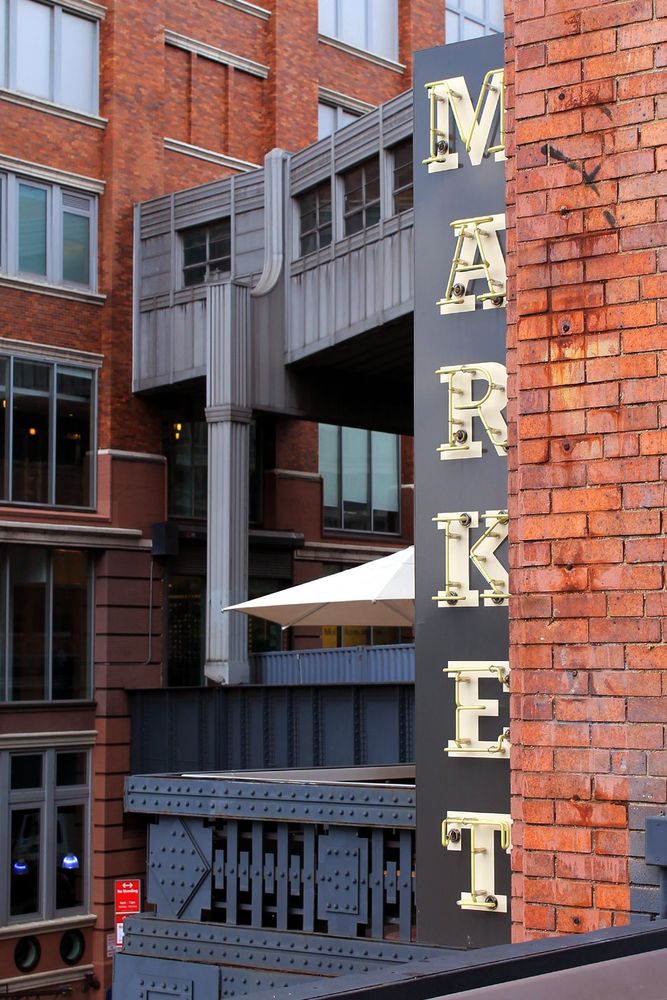 On the High Line park in New York City. There are red brick buildings in both the foreground and the background. The image focuses on a vertical sign in the front-right of the frame that reads Market in yellow neon letters.