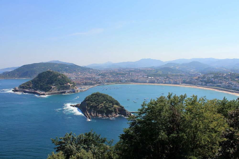 Monte Igueldo in San Sebastian, Spain. This view is from above, accessible by a funicular. In the foreground are trees and the round bay of San Sebastian is visible, lined by the beach. Homes with red tile roofs can be seen in the background on the hills. The trees are a vibrant green and the water is a rich blue.