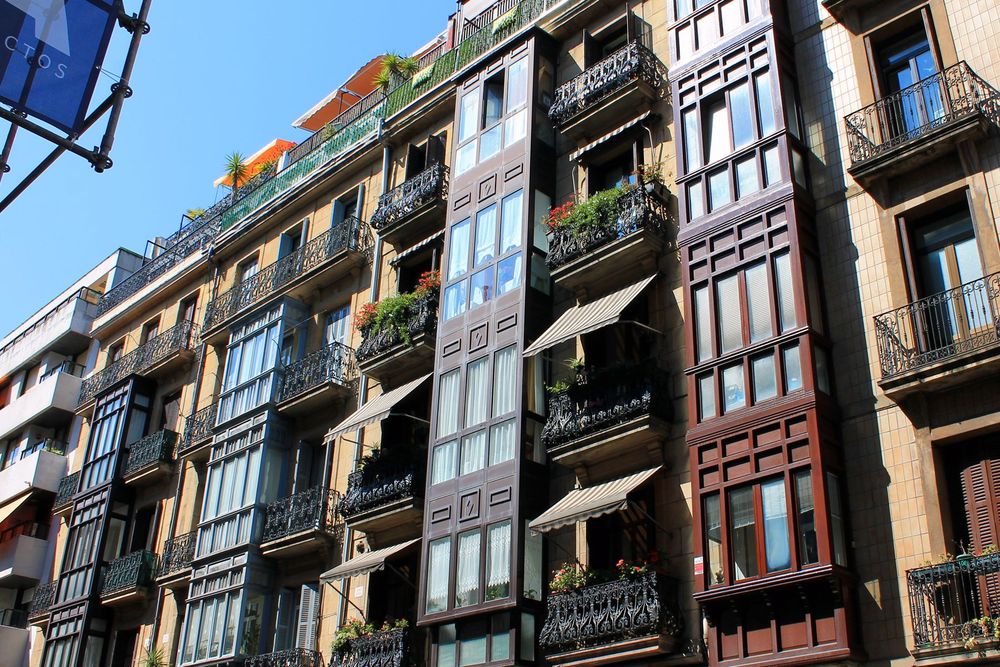 A street in San Sebastian, Spain showing a group of apartments. The building has beige tiles and wooden frames around vertical windows. Each apartment has a black, wrought iron balcony. Many have striped awnings and potted plants.