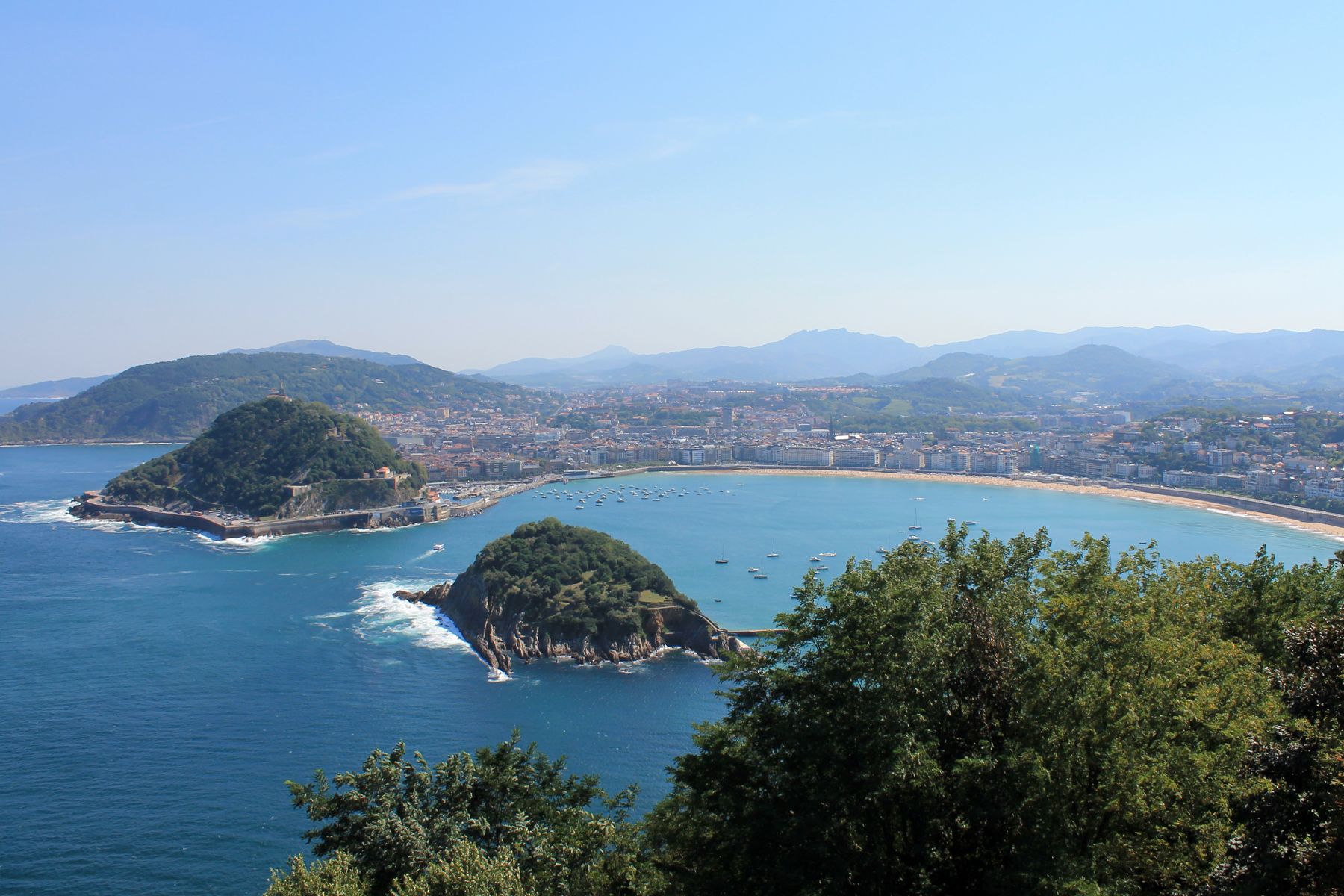 Monte Igueldo in San Sebastian, Spain. This view is from above, accessible by a funicular. In the foreground are trees and the round bay of San Sebastian is visible, lined by the beach. Homes with red tile roofs can be seen in the background on the hills. The trees are a vibrant green and the water is a rich blue.