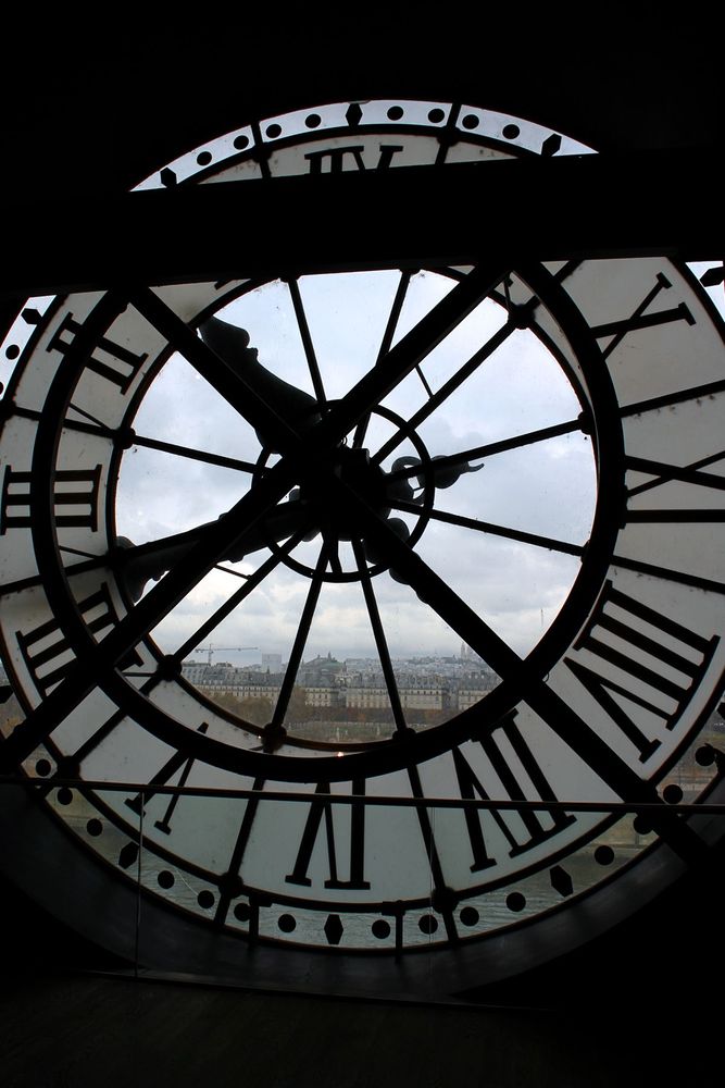 The view from behind a large clock in the Musée d'Orsay in Paris. The clock has large black, metal hands and roman numerals to indicate the hours. The Seine and the streets of Paris can be seen behind the glass. The day outside is grey and overcast.