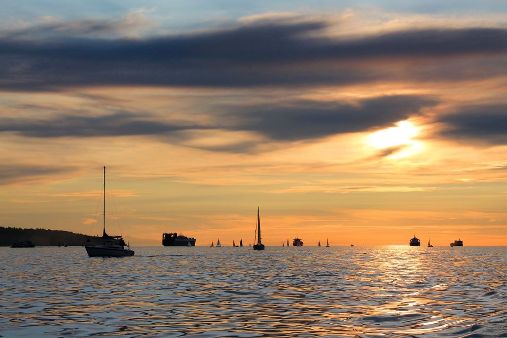 The sun is setting in English Bay, Vancouver and there are some wisps of clouds in the sky. The sky is a mix of yellows, oranges and blues. The water is full of small waves and reflects the colours of the sky. Sailboats are anchored in the foreground and cargo ships can be seen further out. The ships are in silhouette against the setting sun.