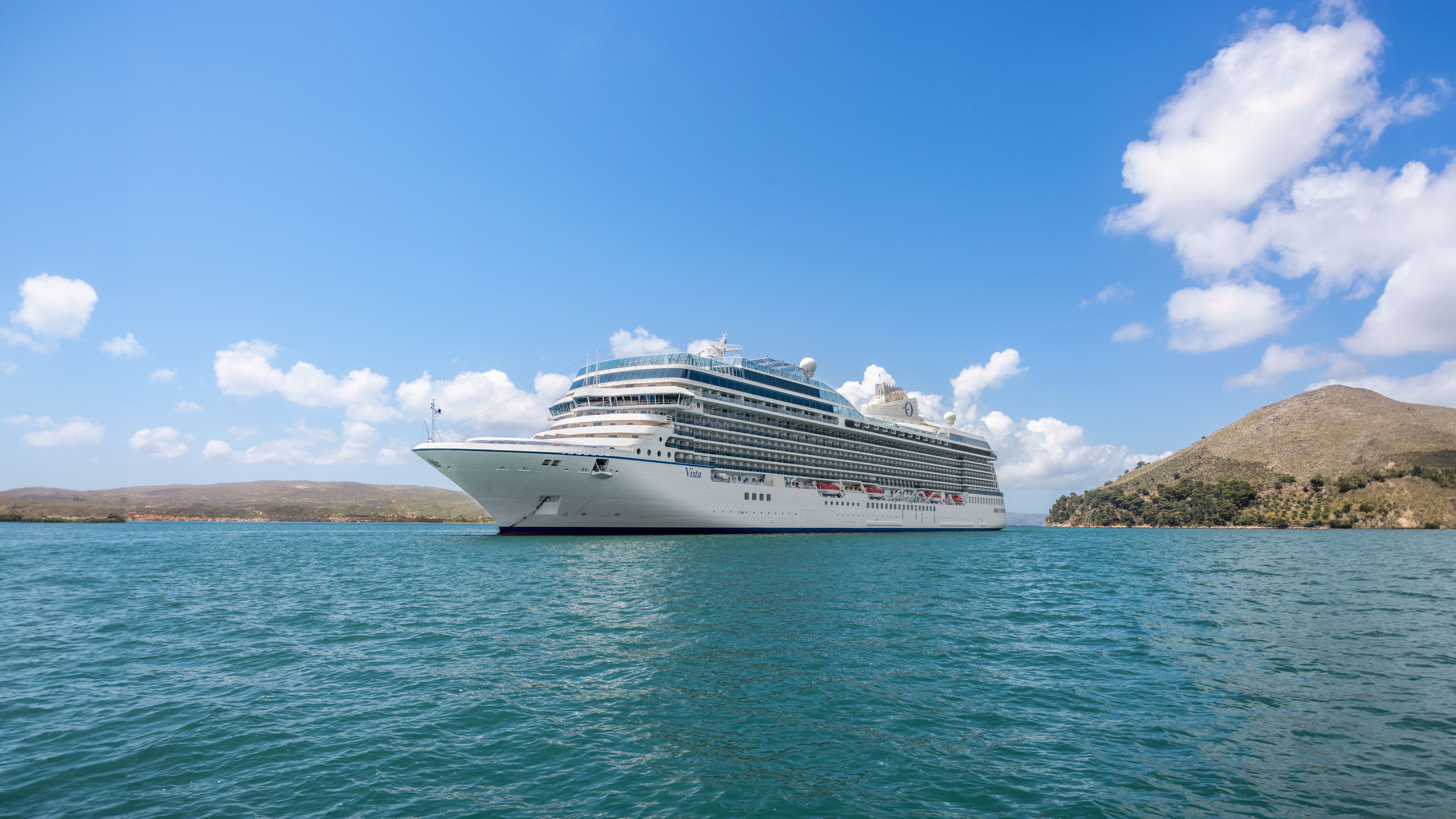 Oceania Cruises' Allura ship majestically cruising in tropical waters with a clear sky and rolling hills in the background.