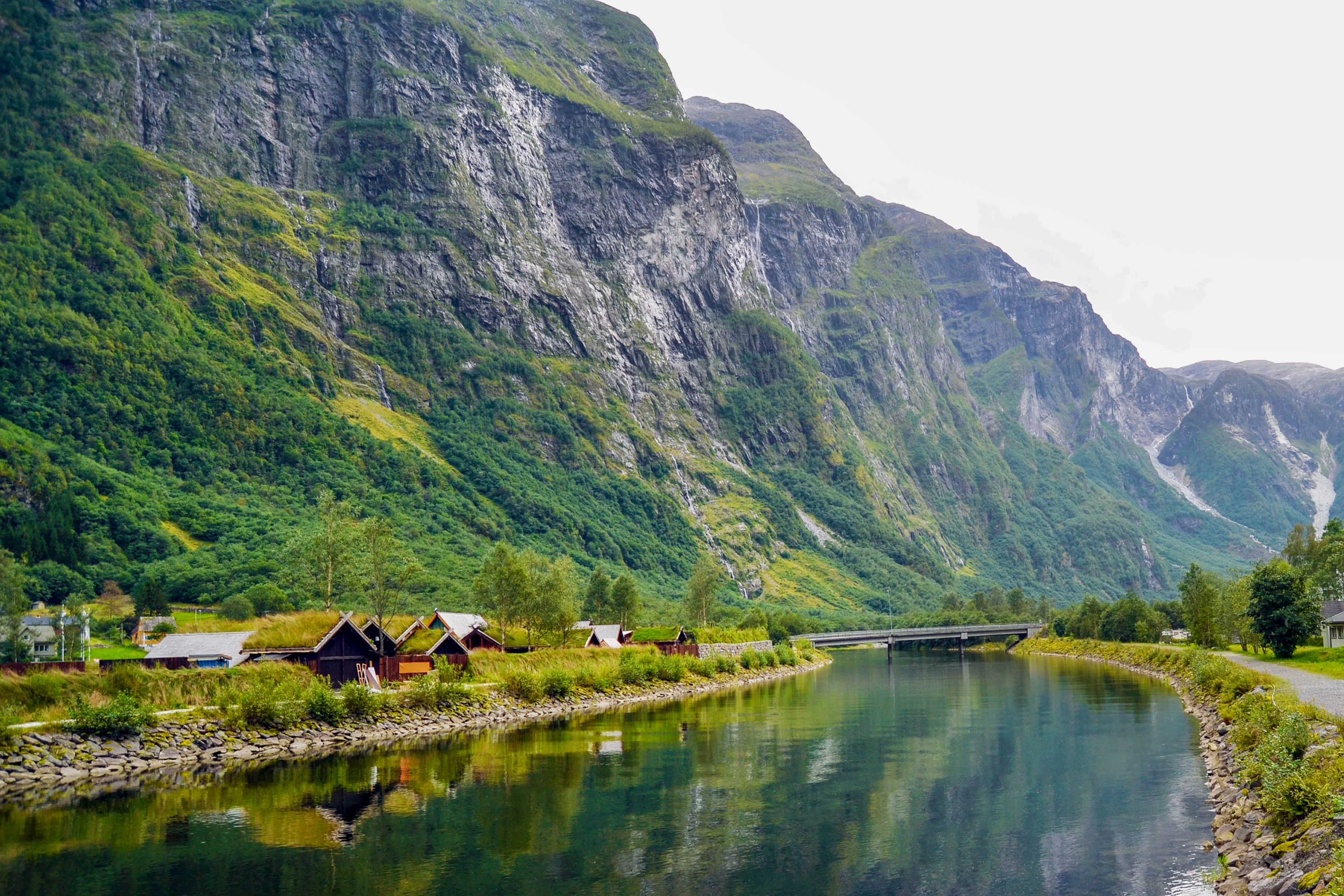 Traditional Norwegian Viking houses with grass-covered roofs nestled at the base of towering green cliffs by a calm river in a fjord.