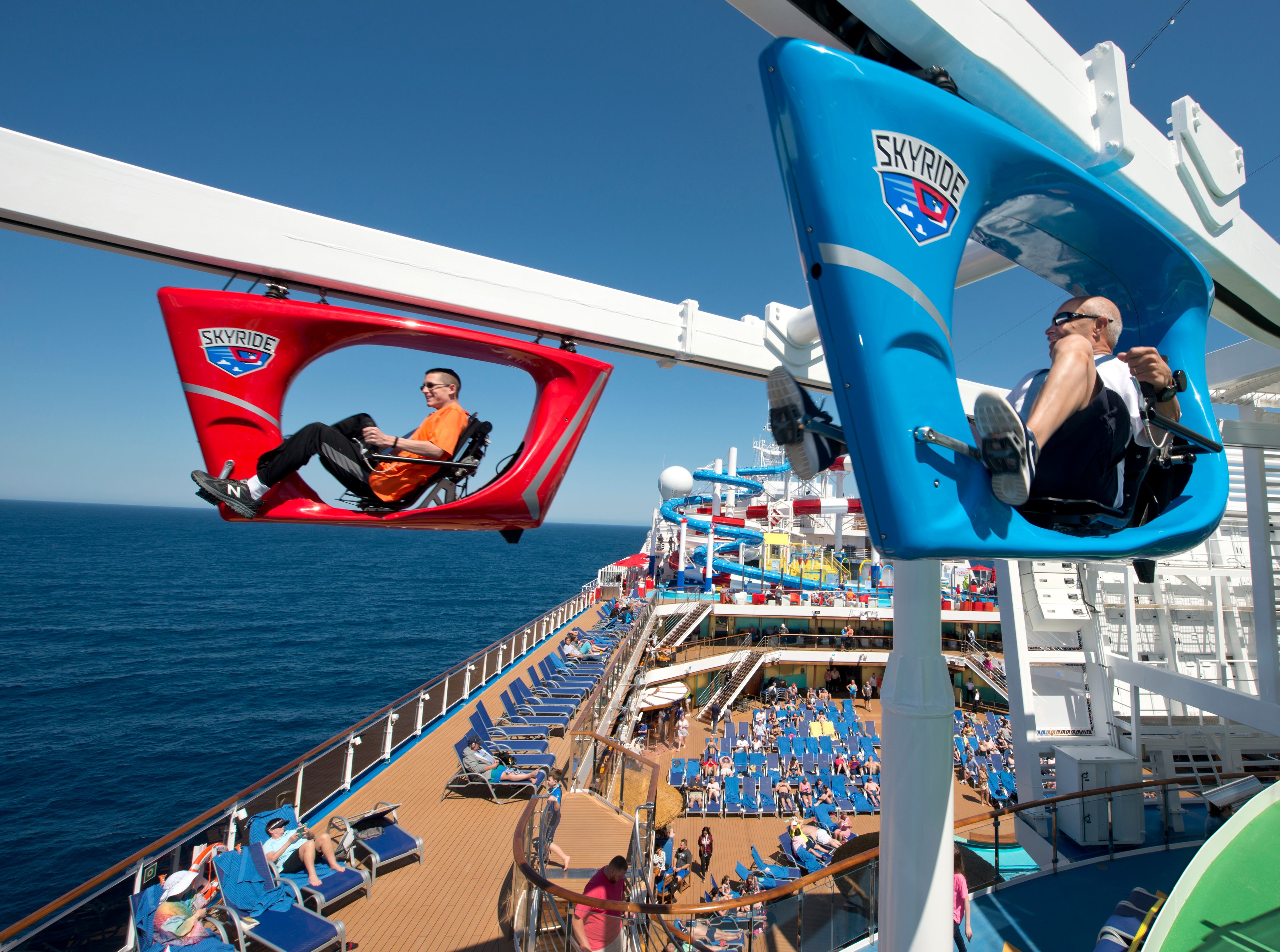 Two passengers riding the Skyride, a suspended bike experience, over the deck of a Carnival cruise ship, enjoying breathtaking ocean views.