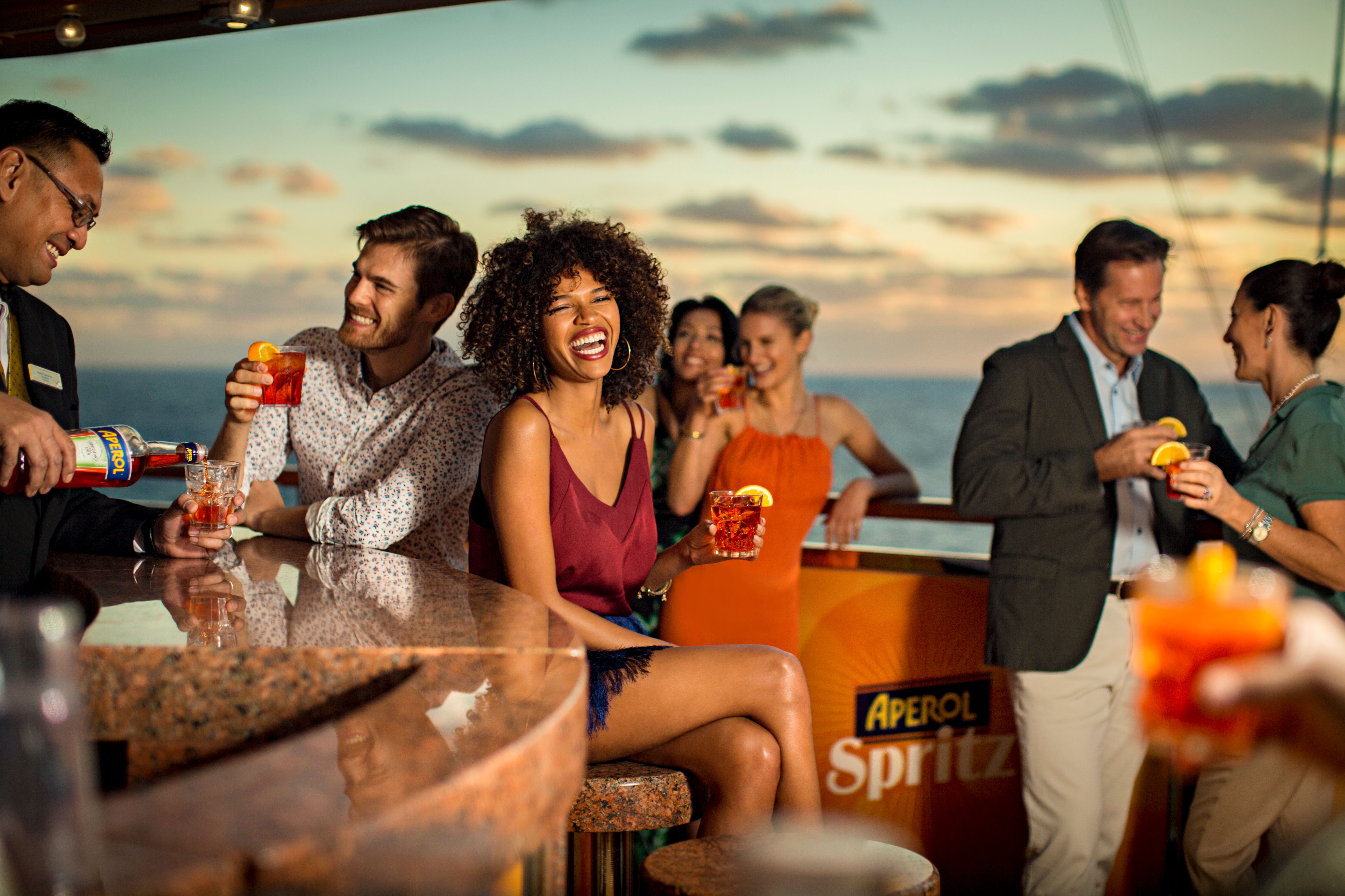 Passengers enjoying lively conversation and refreshing Aperol Spritz at the ship's bar, with a picturesque ocean sunset backdrop.