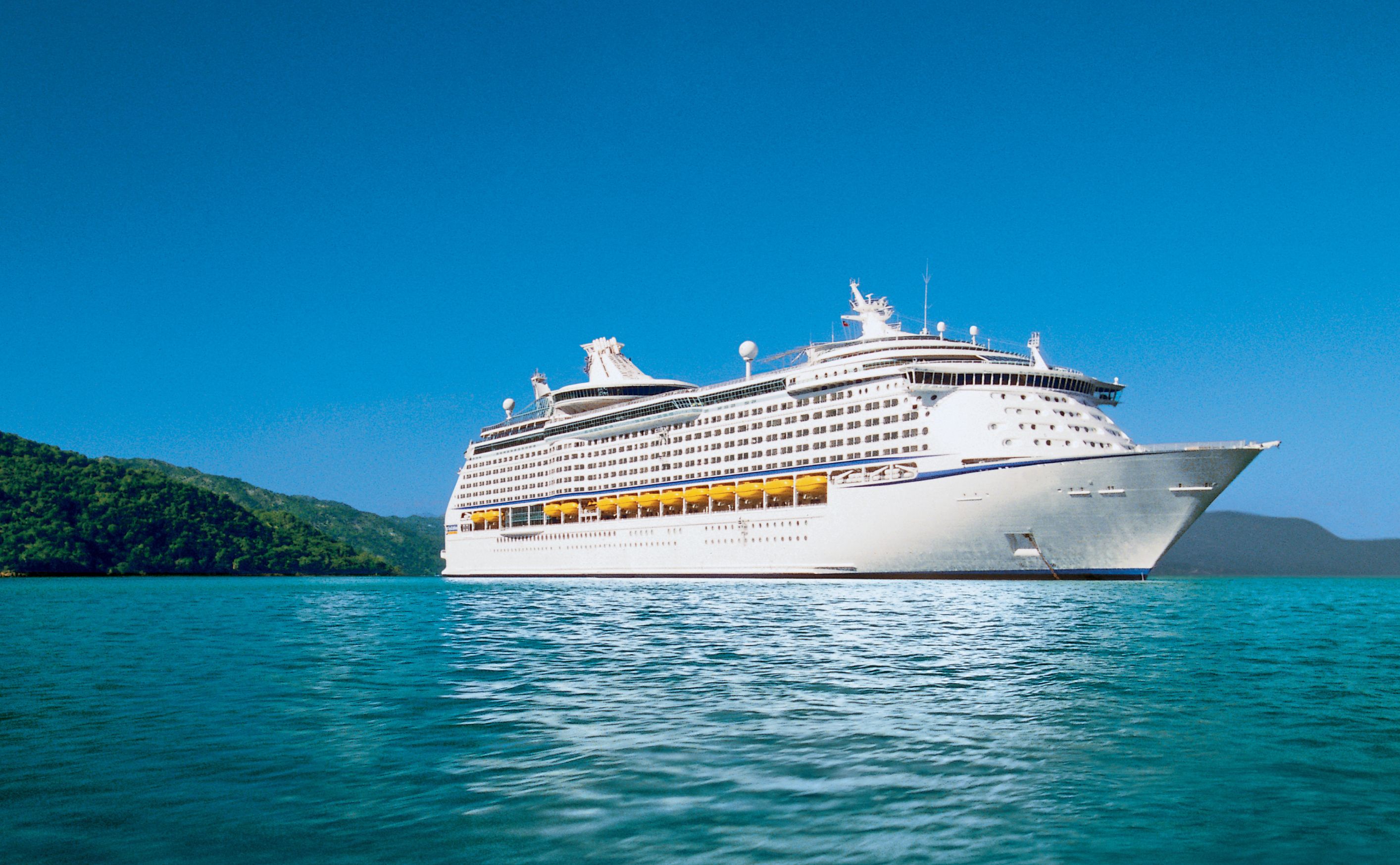 Royal Caribbean's Adventure of the Seas cruise ship anchored in a serene bay with lush green hills in the background, clear blue skies overhead.
