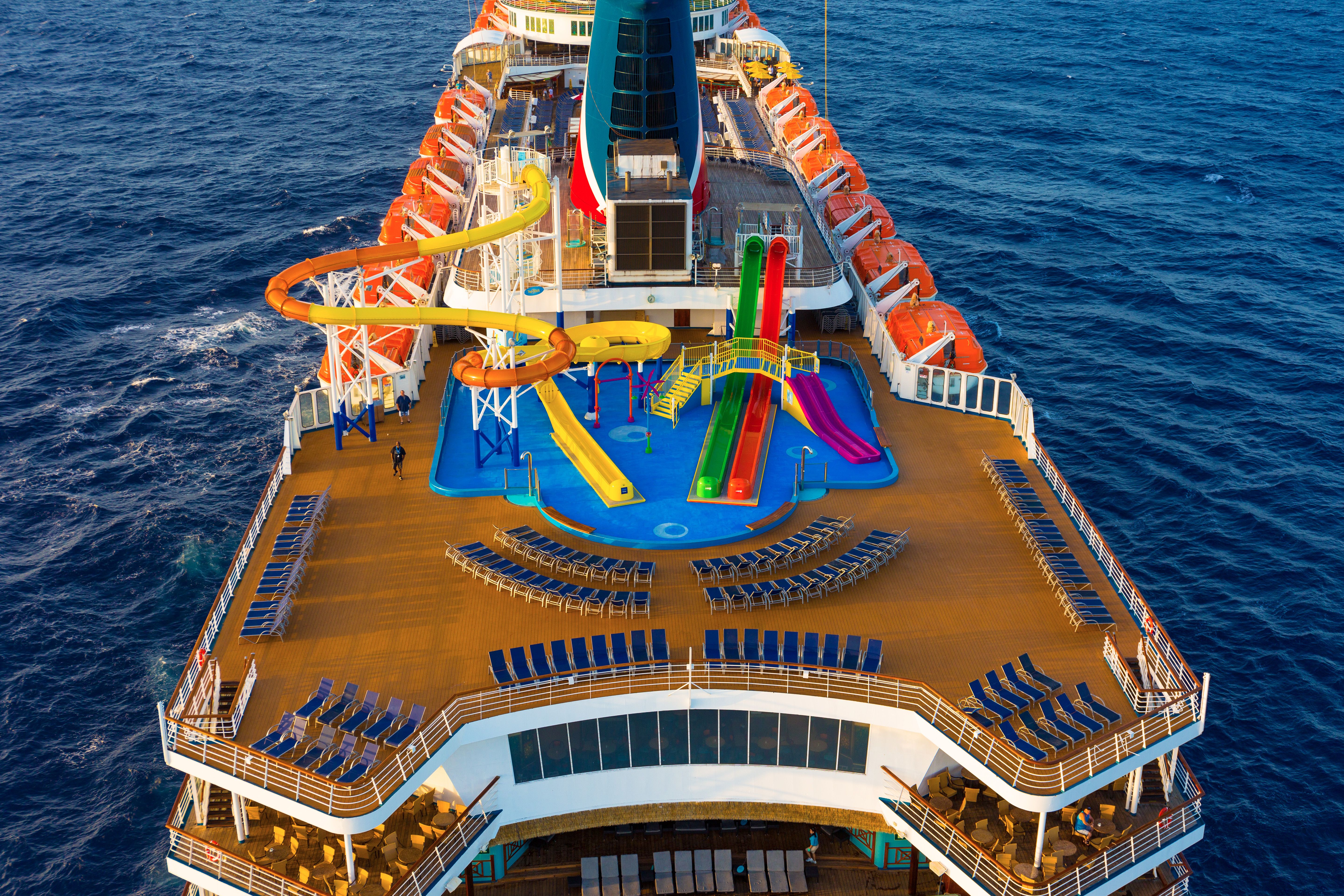 Aerial shot of Carnival Elation's aft deck with colorful water slides, rows of loungers, and the wake of the ship in the ocean.