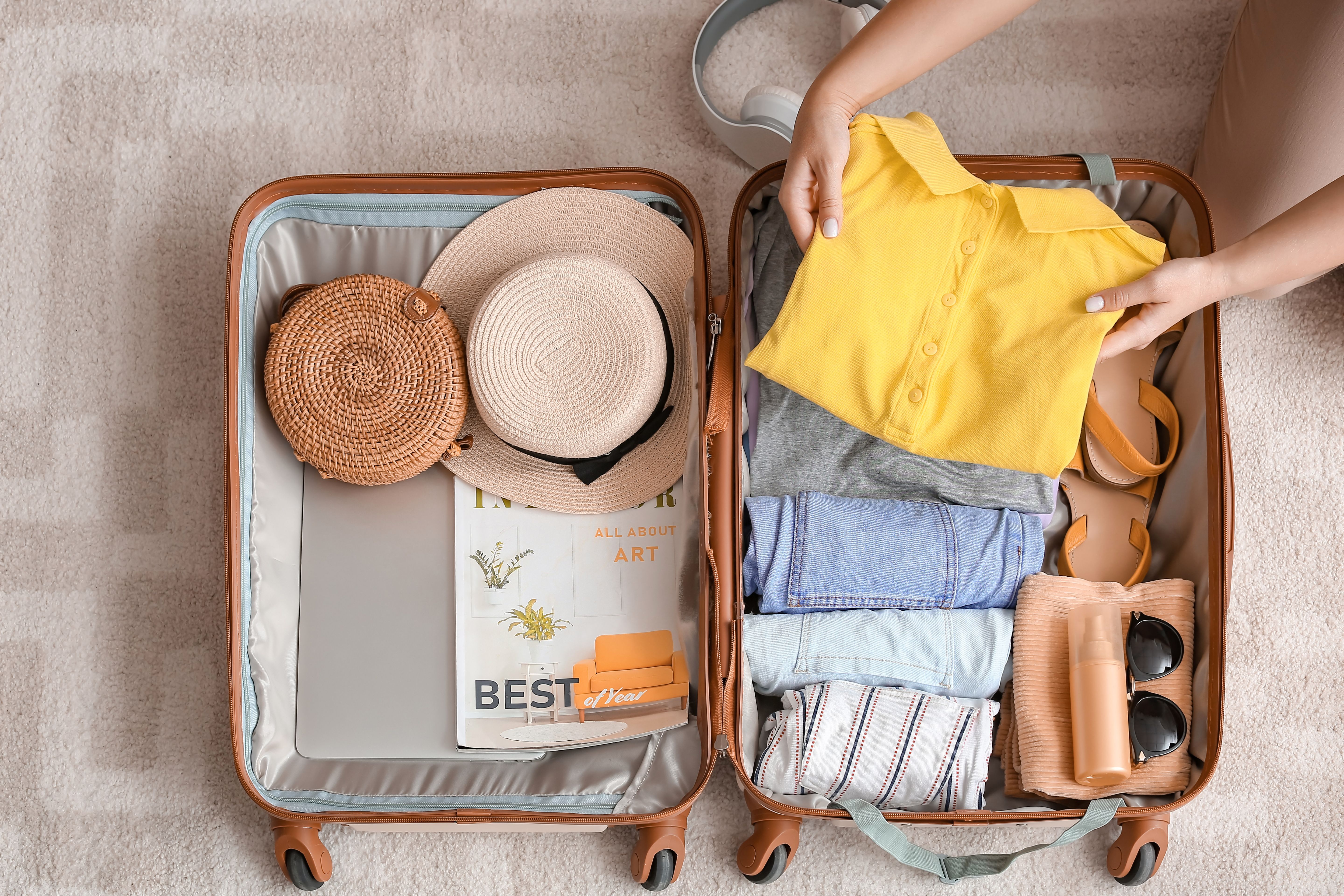 Open suitcase on the floor with neatly packed travel essentials: clothing, a sun hat, art magazine, and toiletries, indicating an upcoming journey.