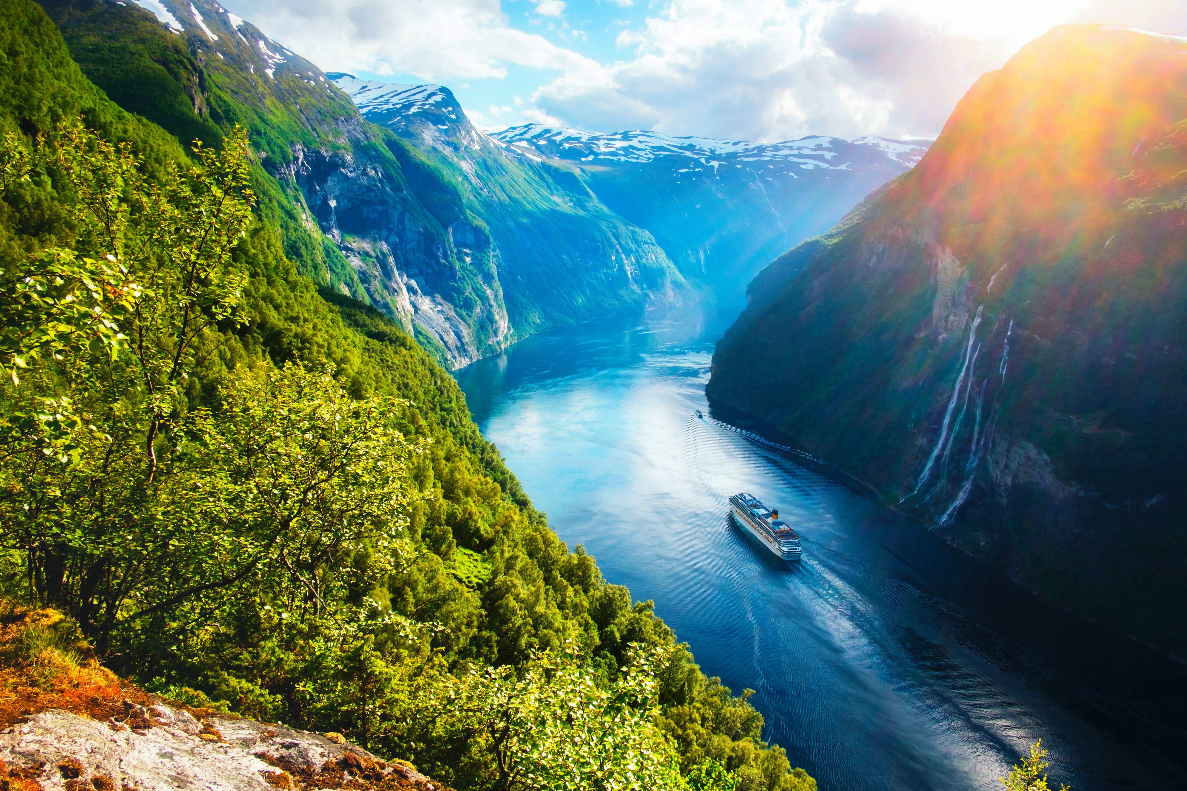 Cruise ship sails through Norwegian fjord, with sunlight filtering through lush mountains.