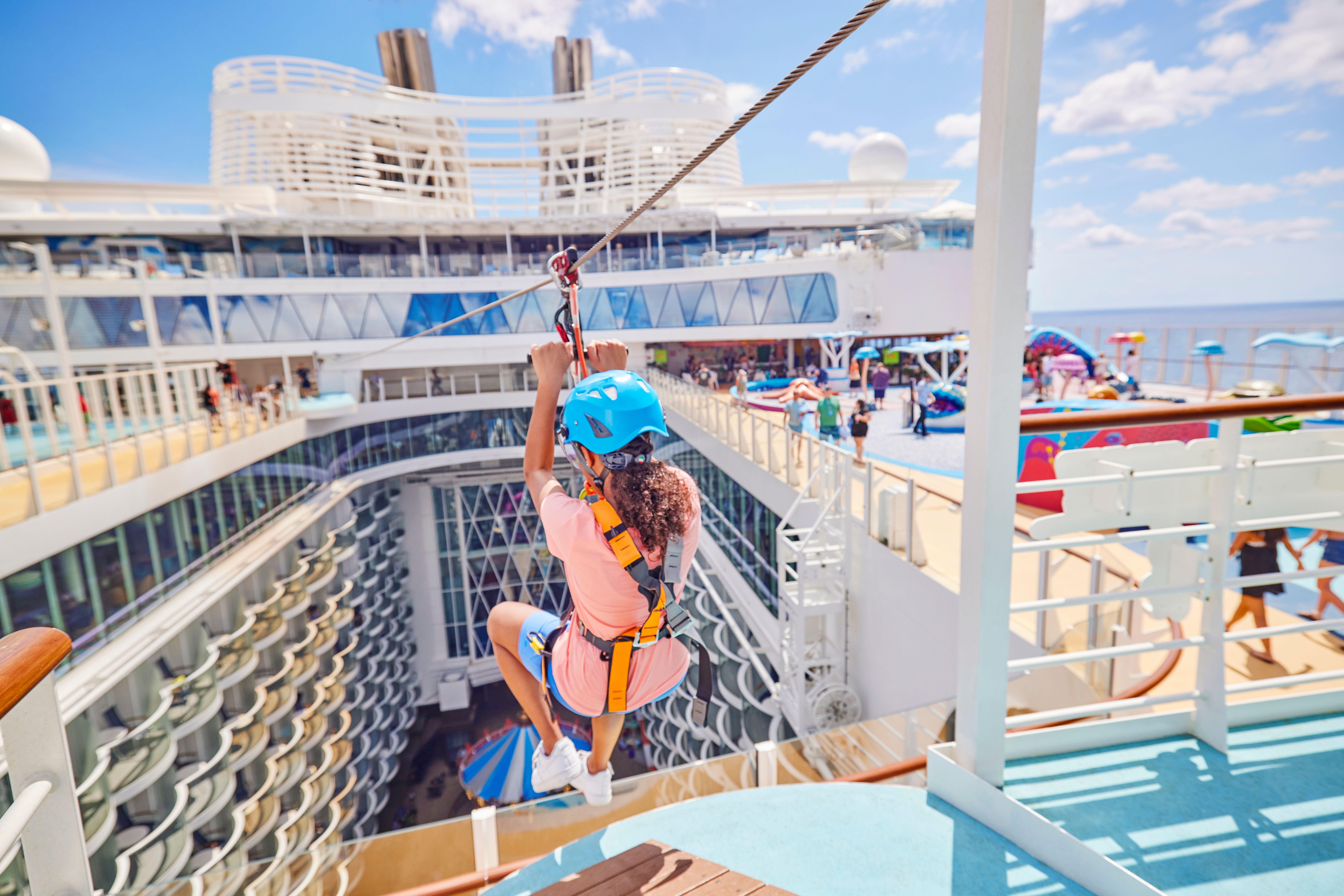 Guest enjoys ziplining on Royal Caribbean's Allure of the Seas with a bustling pool deck and blue skies in the background.