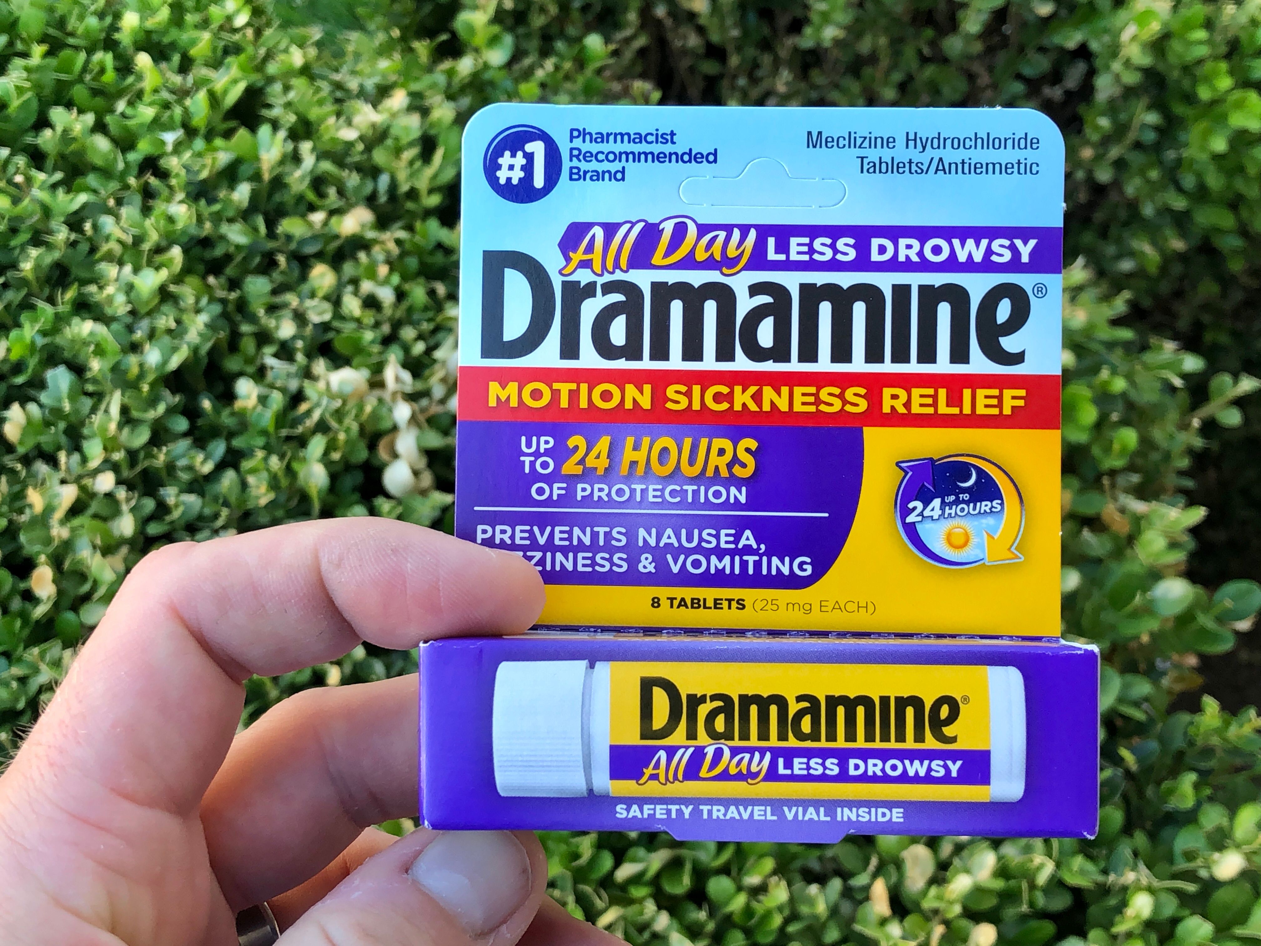 Hand holding a pack of Dramamine against a green hedge, highlighting 24-hour motion sickness relief for cruises.