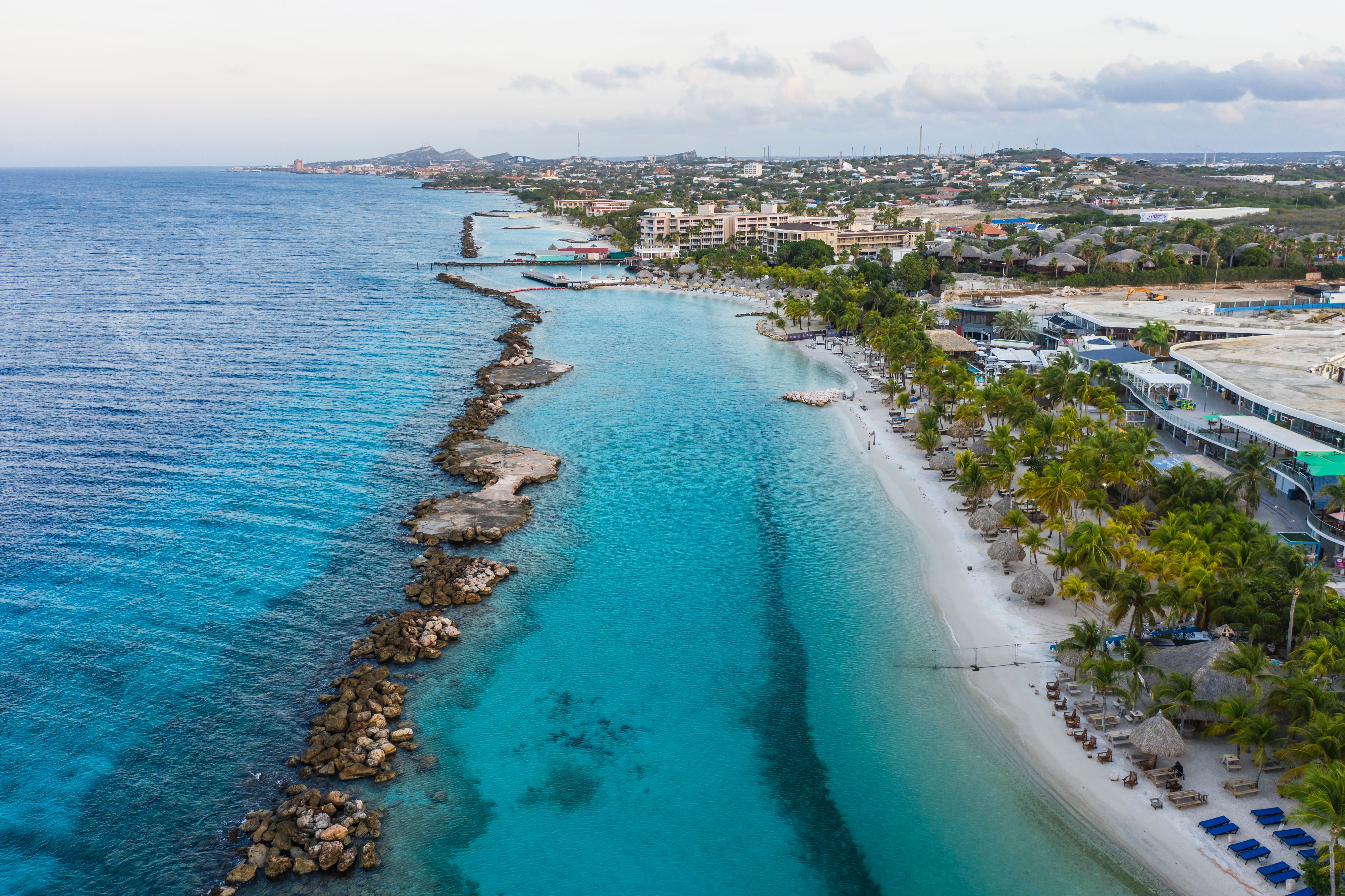Aerial view of Mambo Beach near the cruise port in Curacao, featuring a turquoise sea, a rocky shoreline, and a series of beachfront properties and palm trees.