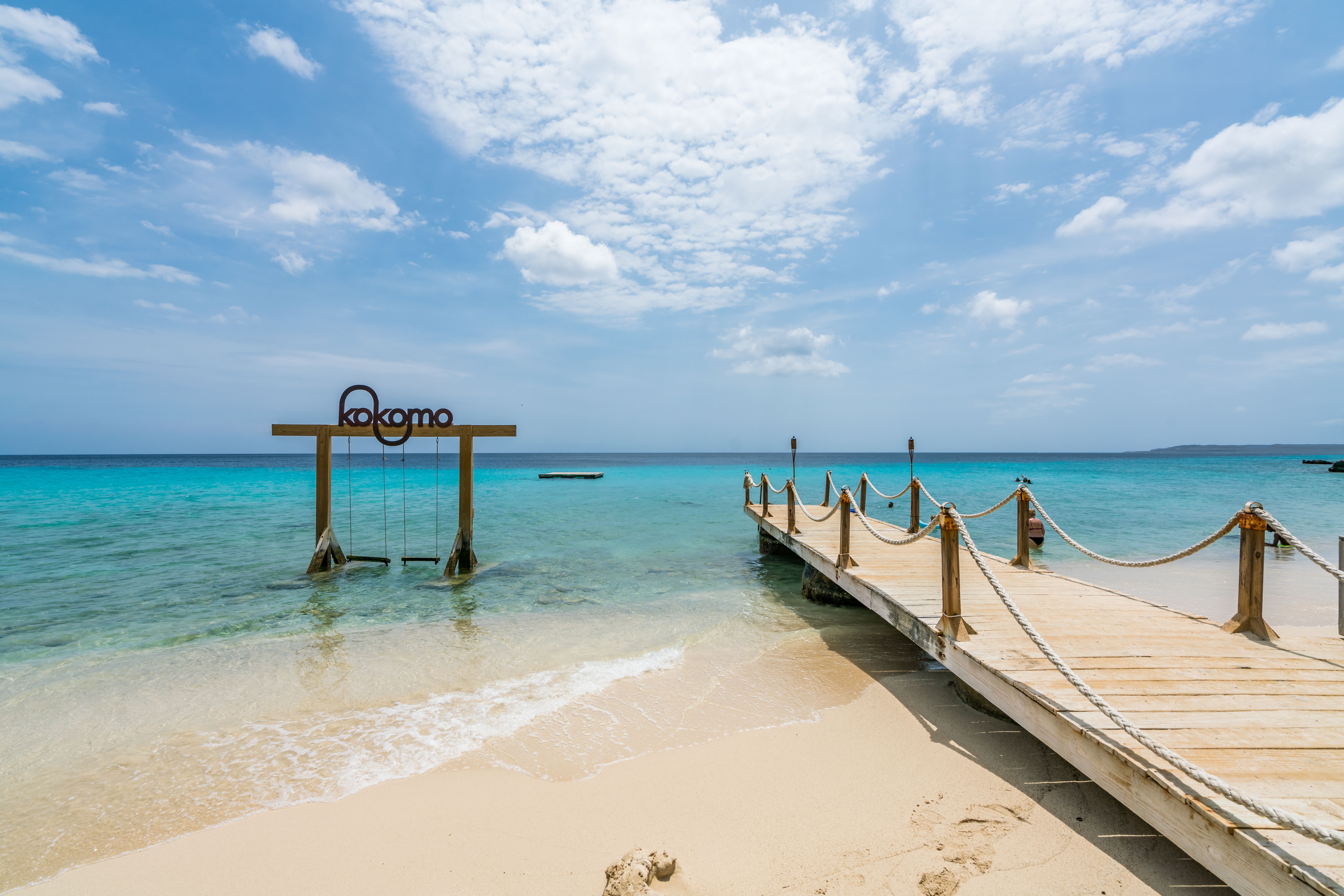 Serene view of Kokomo Beach in Curacao with a wooden pier and rope swings under a sign, white sandy shore, and crystal-clear turquoise waters.
